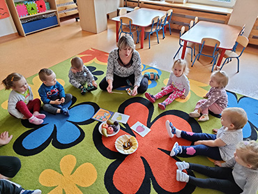 Na drugim zdjęciu widać grupę dzieci, które siedzą na dywanie w trakcie zajęć o warzywach i owocach.