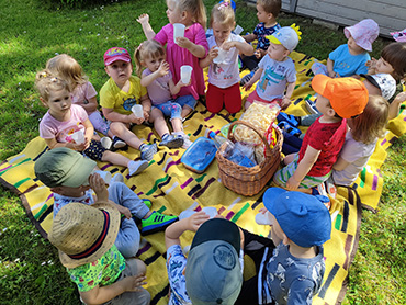 Zdjęcie przedstawia całą grupę dzieci, które siedzą na kocyku podczas pikniku.