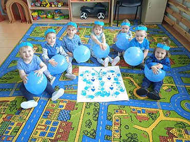 Dzieci siedzące w półkolu ubrane na niebiesko, trzymające niebieskie balony. Po środku leży praca którą wykonały dzieci.