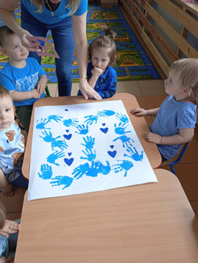 Dzieci siedzące przy stoliku. Na stole leży praca niebieskiego motyla z odbitych przez dzieci rączek.