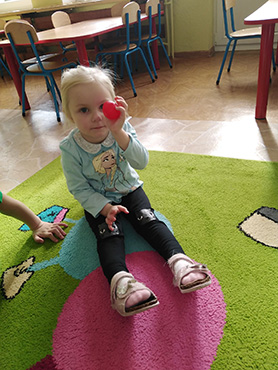 Dziewczynka siedzi na dywanie , a w rączce trzyma czerwone jabłko 